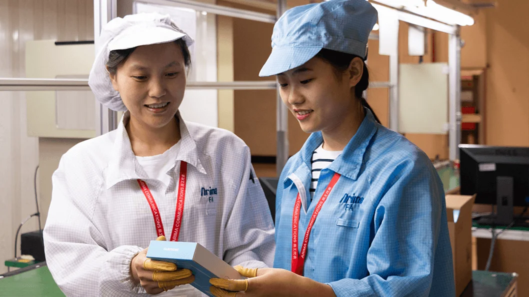 Two Asian women in a manufacturing setting, examining a Fairphone package. The woman on the left, in a white lab coat and cap, holds the box, while the woman on the right, in a blue work shirt and cap, looks on attentively. They are in a facility with industrial shelving in the background, suggesting a focus on ethical or sustainable electronics production.