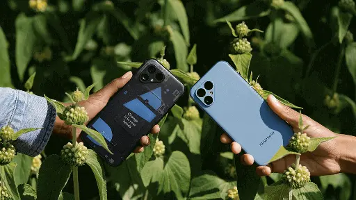 Two hands holding Fairphone 5 smartphones against a background of green leaves and small flower buds. The left hand holds a black Fairphone 5 with a blue accent on the back, while the right hand holds a light blue Fairphone 5.