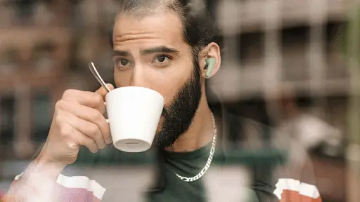 Man with a beard sipping coffee while wearing Fairphone wireless earbuds, captured through a blurred café window.